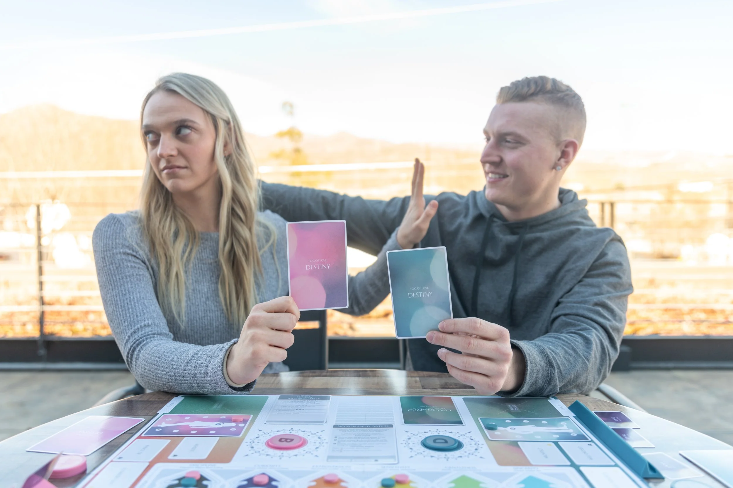 Two people playing a card game outdoors at a table, with game materials and cards in hand, high-five gesture, and a blurred background of a fence and mountains.