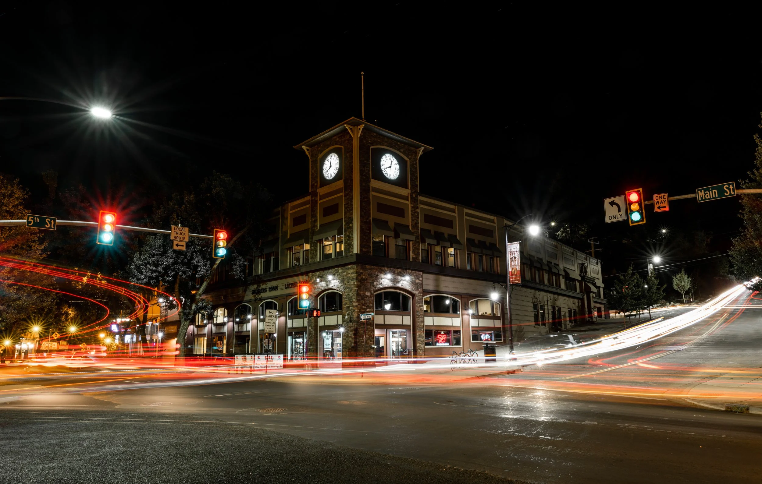 Nighttime city street scene with a historic building featuring a clock tower at the corner of 5th Street and Main Street, with traffic lights and streaks of car lights from long exposure.
