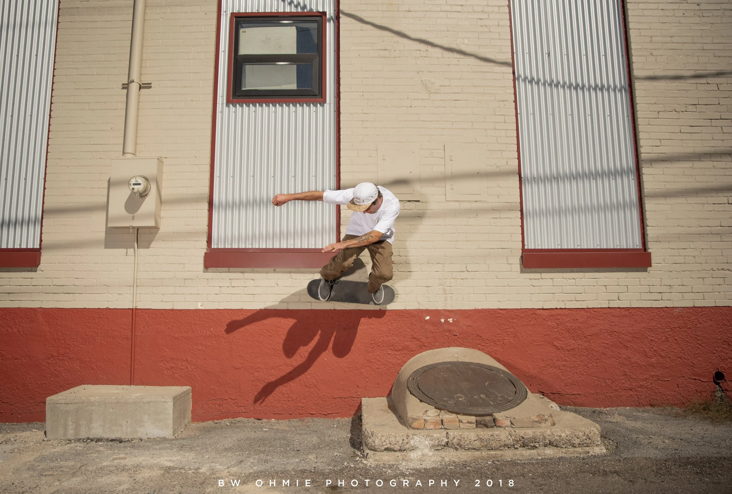 A man skateboarding off a ledge in front of a building with brick and metal siding, with shadows cast on the wall.