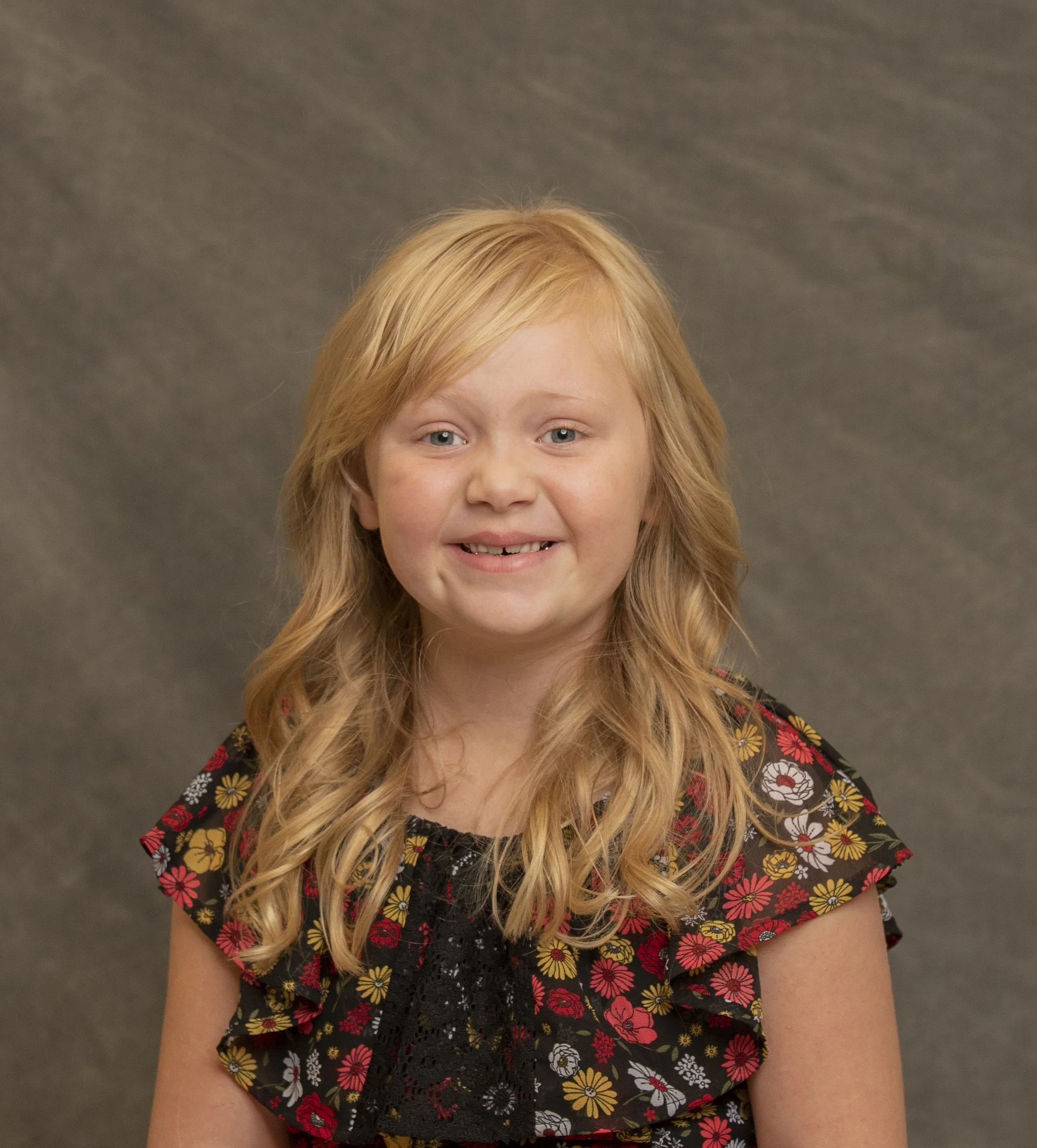 A young girl with long red hair wearing a black floral dress, smiling at the camera against a gray backdrop.