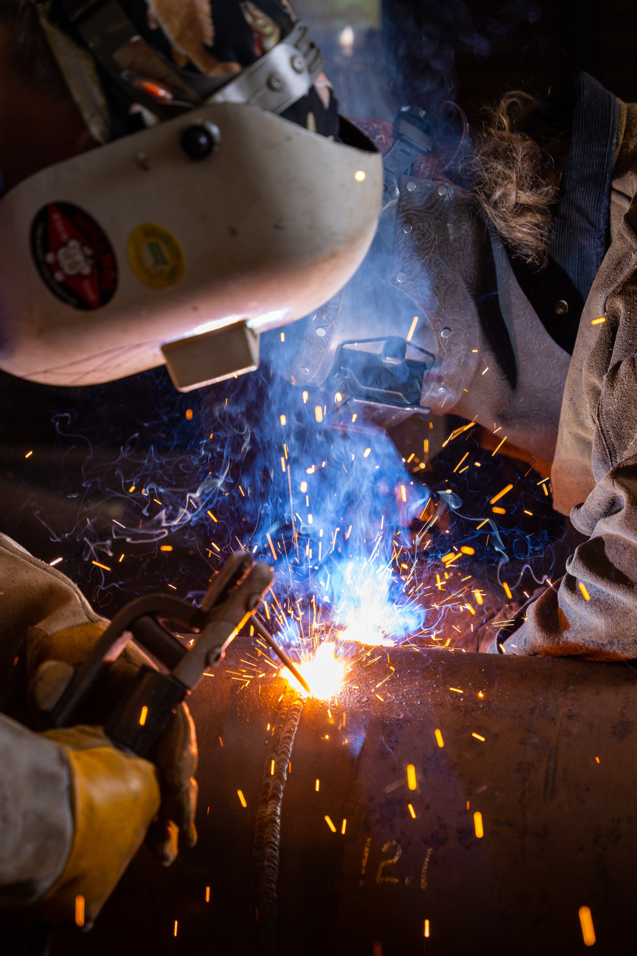 Welder wearing protective gear welding metal with sparks and bright blue flame.