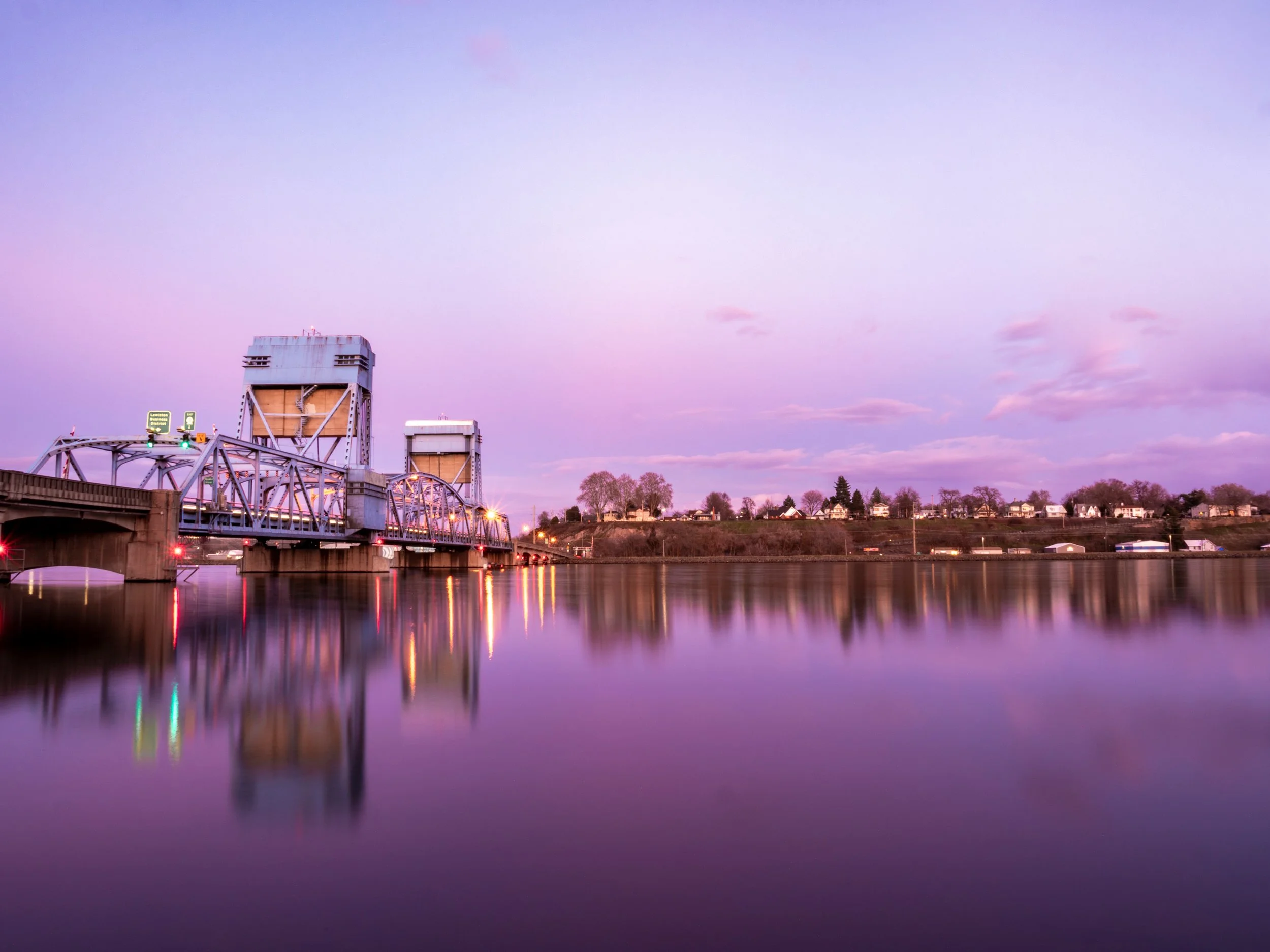 Bridge over a river at sunset with purple sky and residential houses in the background.