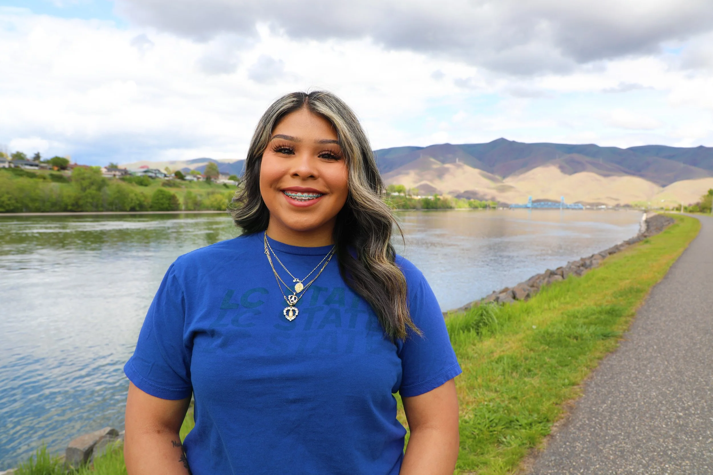 A young woman with long wavy hair, wearing a blue T-shirt, jewelry, and braces, smiling while standing outdoors near a river with a rocky bank, green grass, trees, a bridge, and mountains in the background on a partly cloudy day.