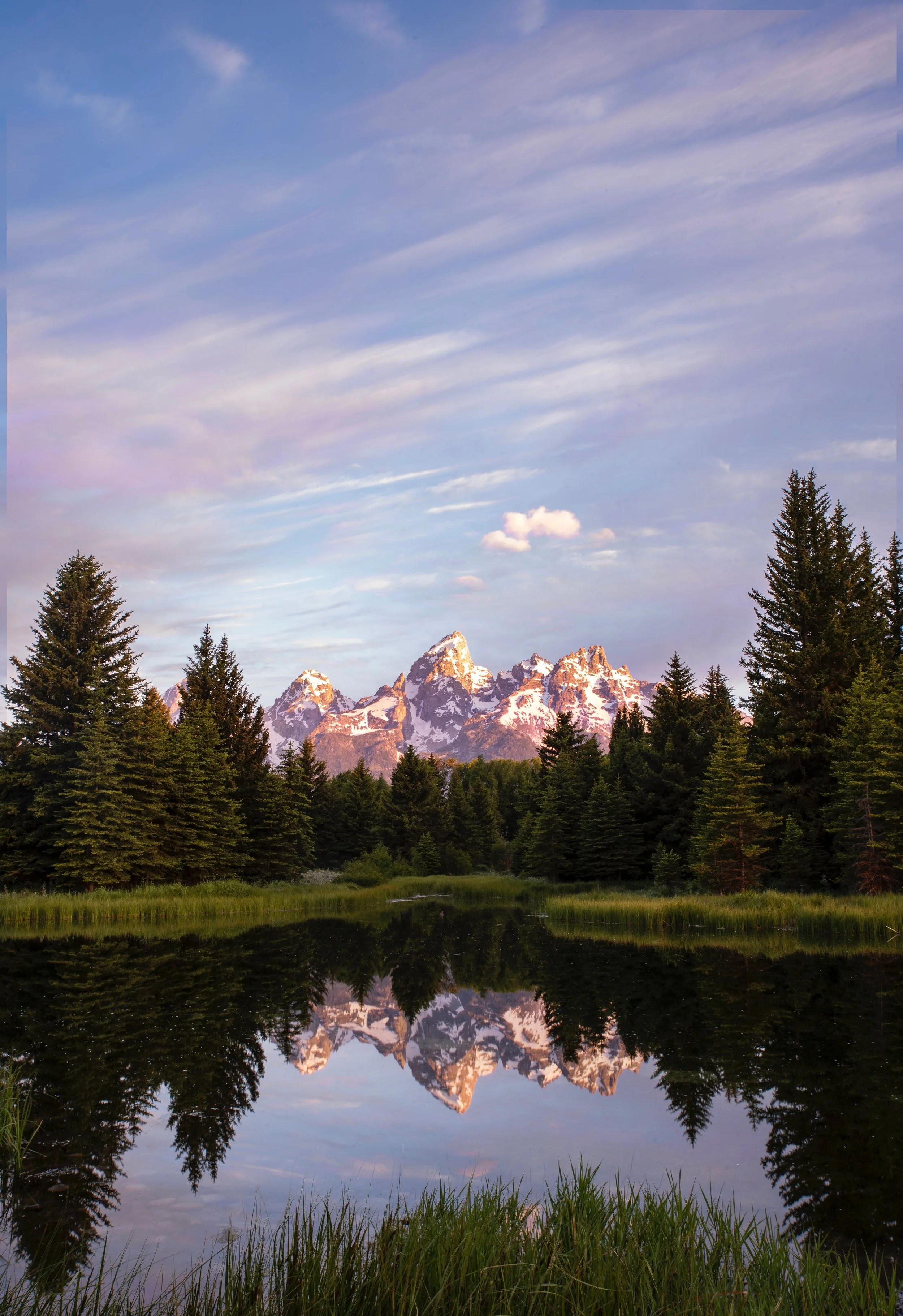 Snow-capped mountains reflecting in a calm lake surrounded by evergreen trees under a partly cloudy sky.