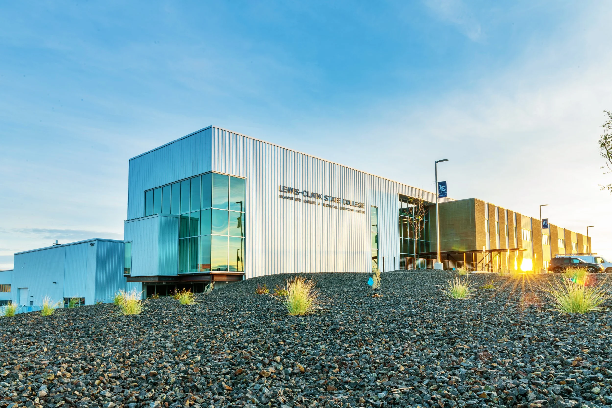 Modern college building with the sign 'Lewis-Clark State College' on the facade, large glass windows, and a parking lot in the foreground during sunset.