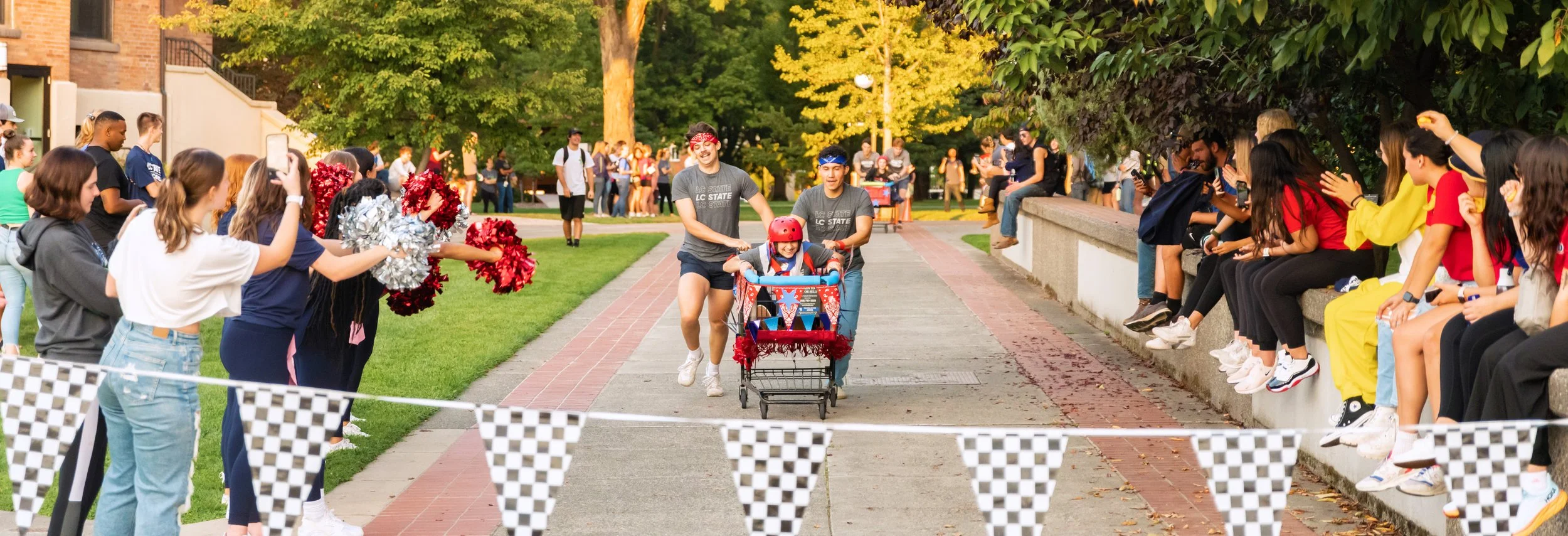 People cheering as a child rides a decorated bicycle in a race, with spectators sitting and standing along the sides and taking pictures, in a park with trees and sunlight.