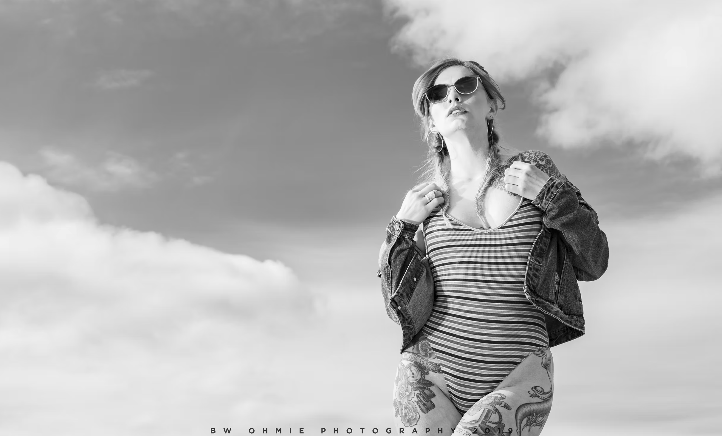 Black and white photo of a woman with tattoos wearing sunglasses and a striped swimsuit, standing outdoors against a cloudy sky backdrop.