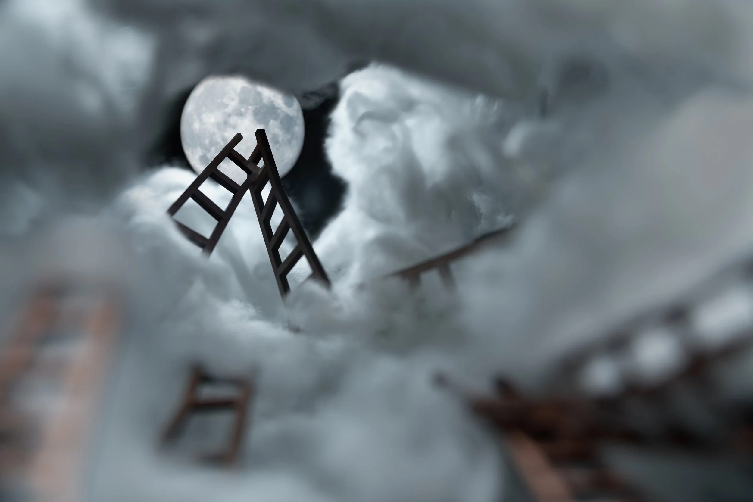 Miniature ladder and staircase leading towards the moon in a cloudy sky, creating an abstract scene.