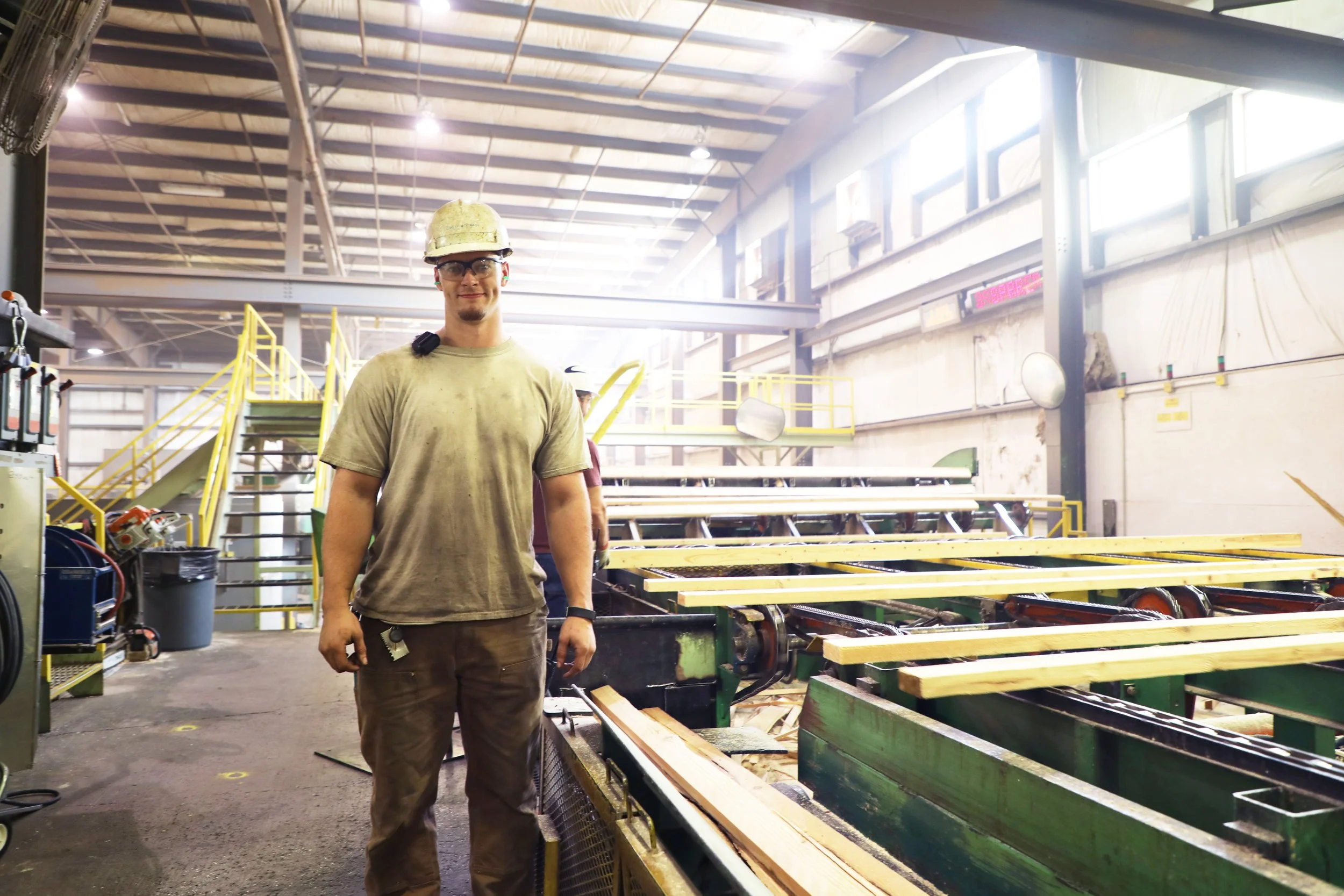 A man in construction gear standing inside an industrial woodworking facility with wood beams and industrial machinery around him.