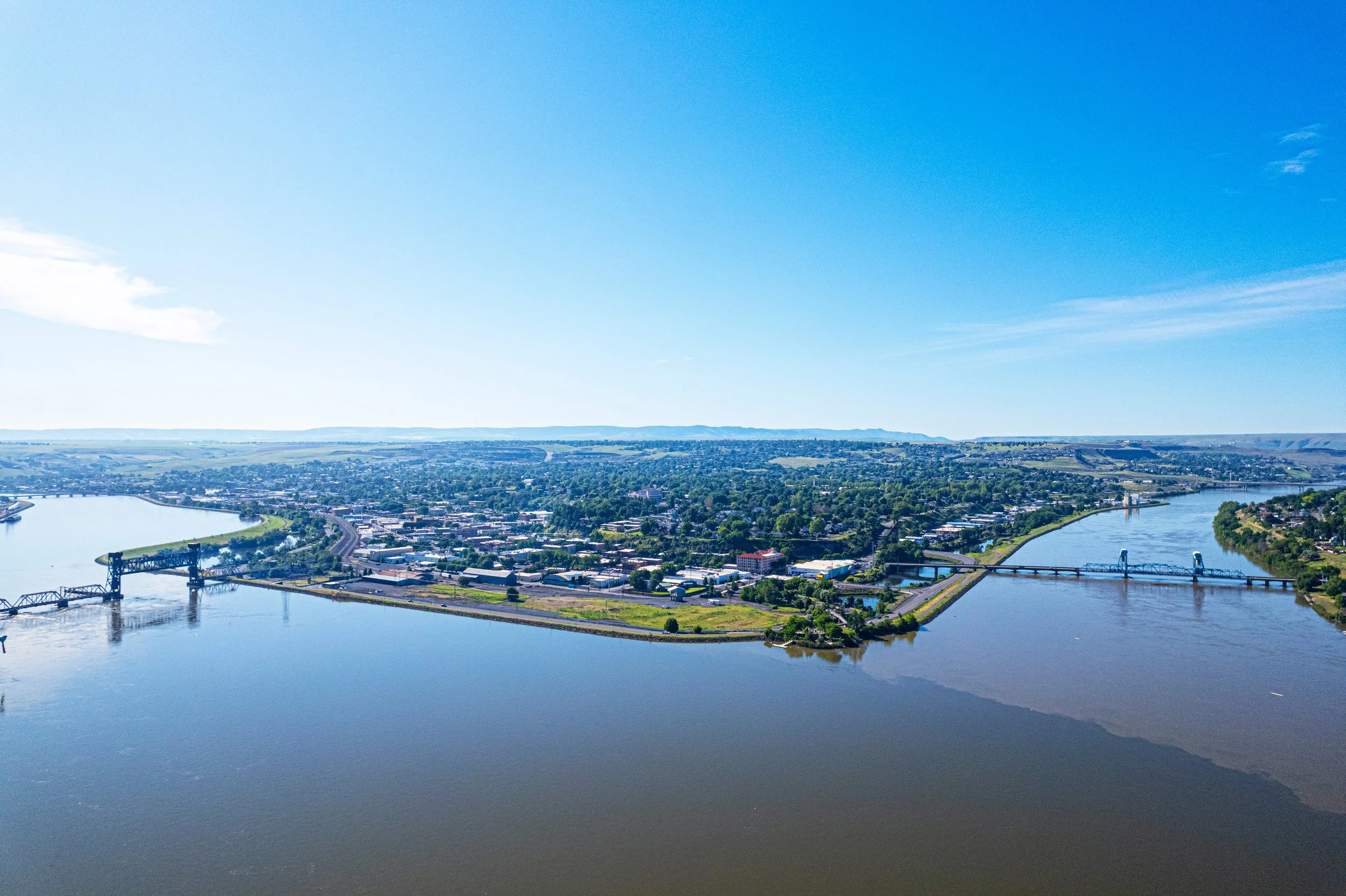 An aerial view of a river with a bridge, a cityscape, and green hills under a clear blue sky.