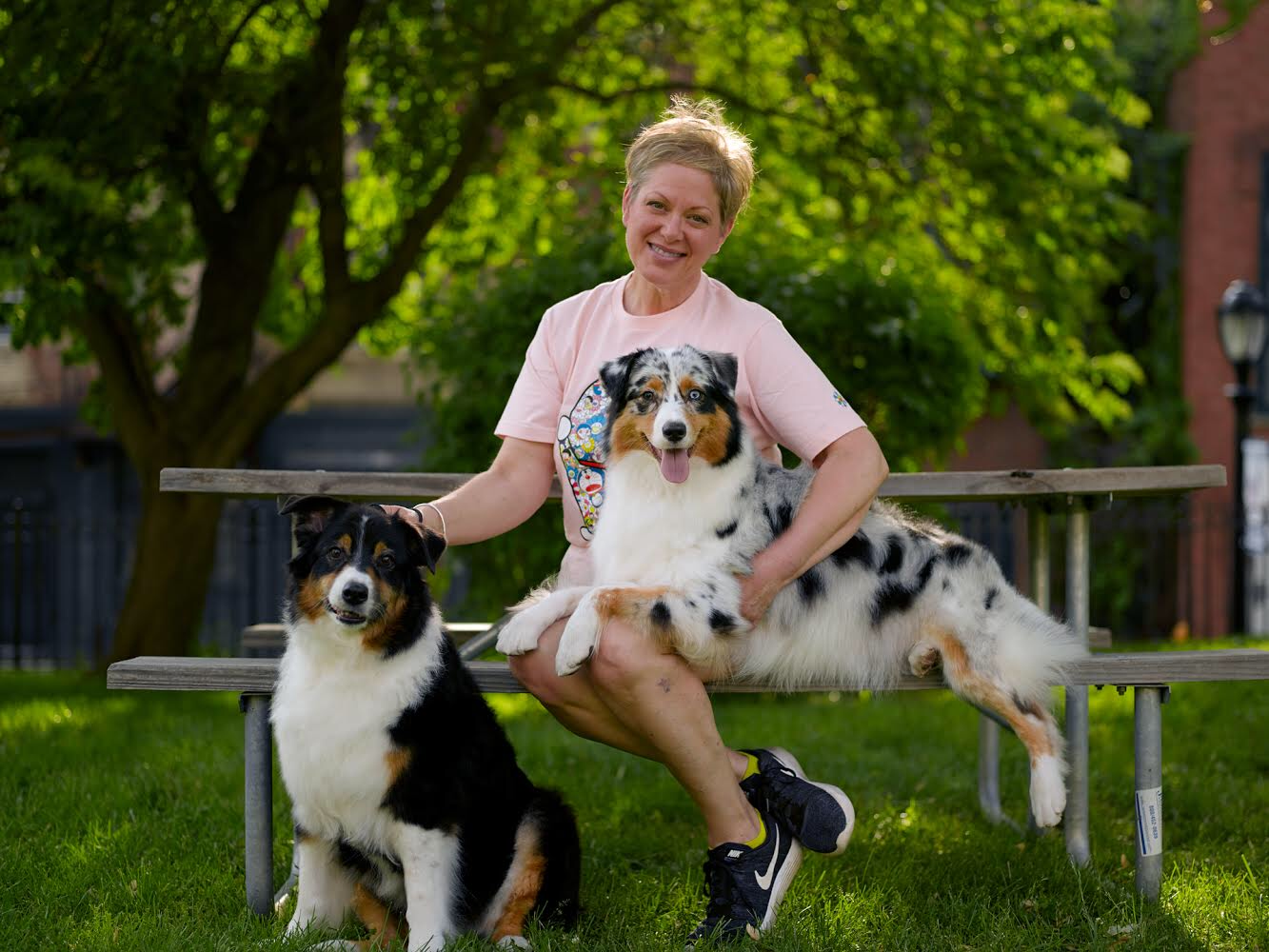 Woman sitting on park bench with two Australian Shepherd dogs, one sitting on the ground, the other lying across her lap, outdoors with green trees and a brick building in the background.