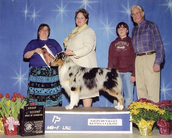 A group of four people and an Australian Shepherd dog at a dog show, with a blue starry backdrop and flowers, the dog is on a platform labeled Saginaw Valley Kennel Club, and there is a black sign indicating second place in the group.