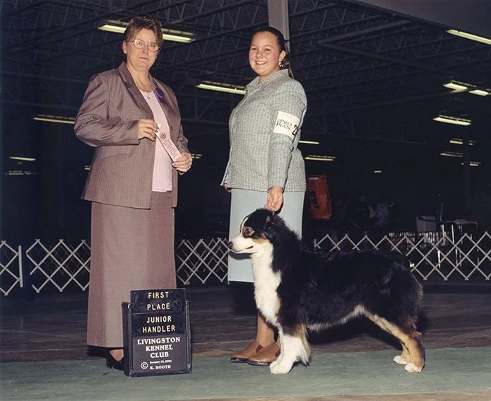 A young woman and an older woman standing with a black and white Australian Shepherd dog at a dog show. The young woman is smiling and holding the dog's leash, while the older woman is holding a ribbon. A sign in front indicates they won first place at the Livingston Kennel Club Junior Handler event.