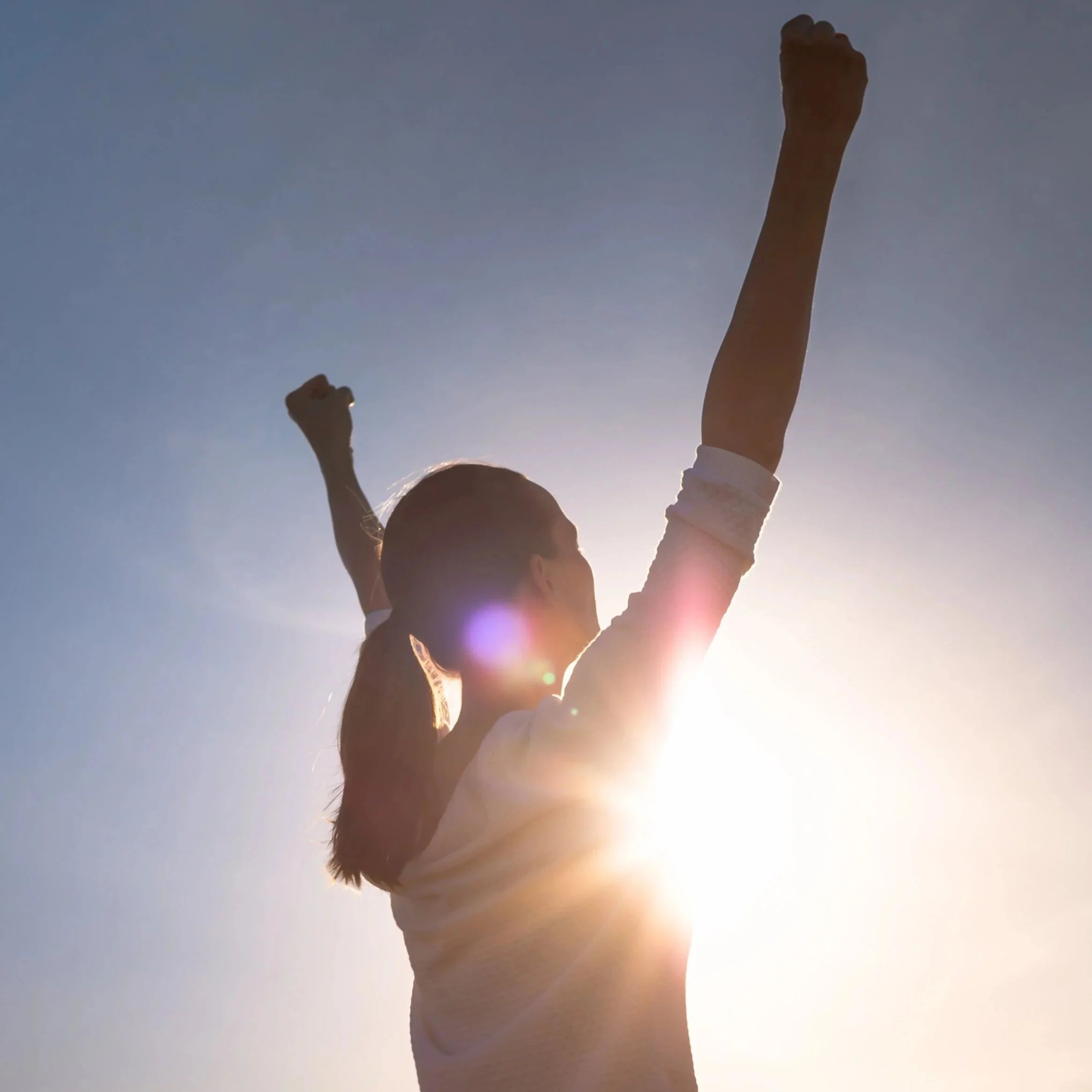 A woman with her hair in a ponytail raising her fists in the air with sunlight shining behind her, celebrating outdoors.