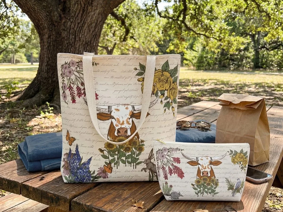 Texas Lone Star Wilds market tote and accessory pouch displayed beneath an oak tree, featuring original longhorn and Texas wildflower artwork.
