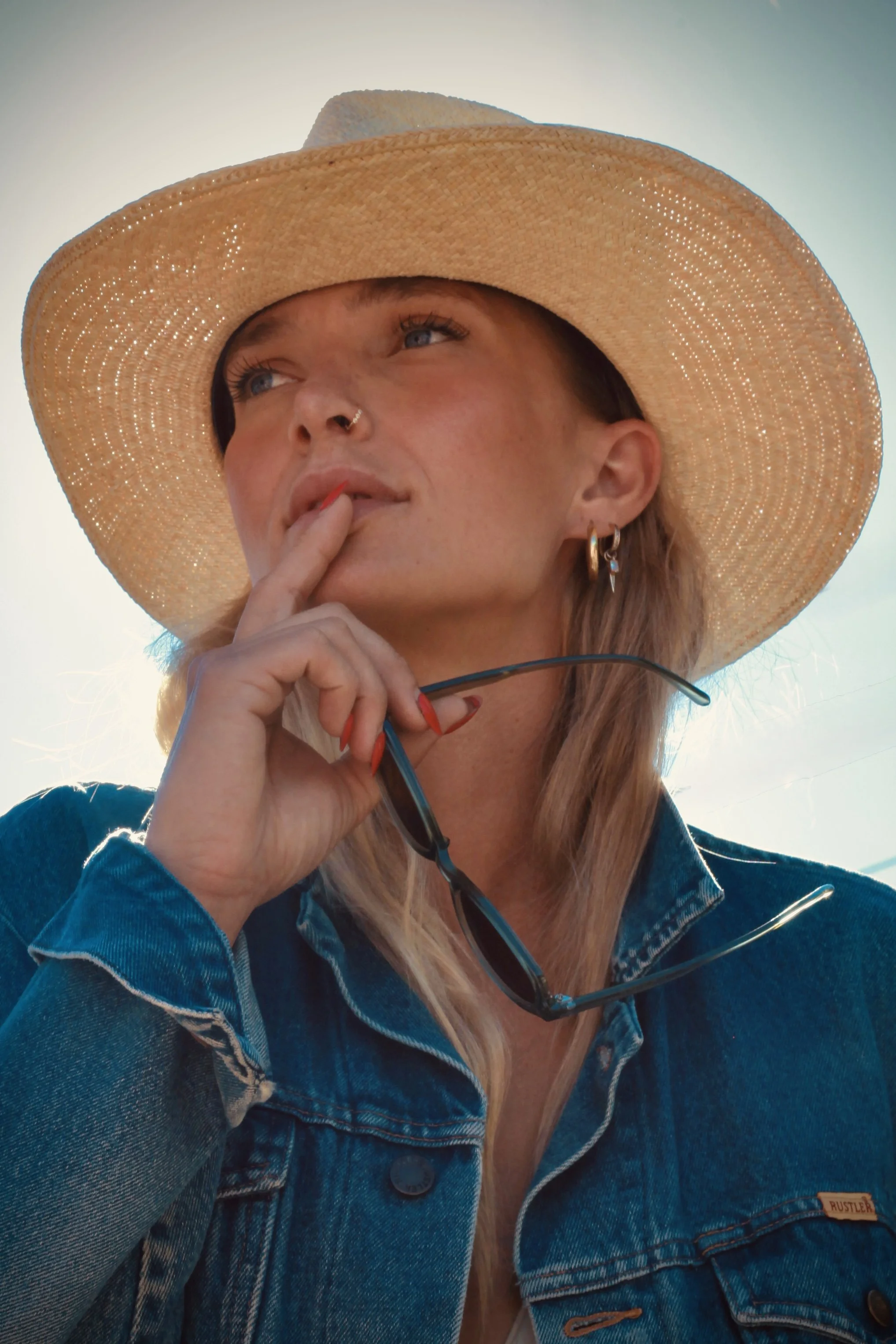 Portrait of a woman with blue eyes, wearing a large straw sun hat, denim jacket, earrings, and a nose ring, holding glasses near her mouth, outdoor setting with sunlight.