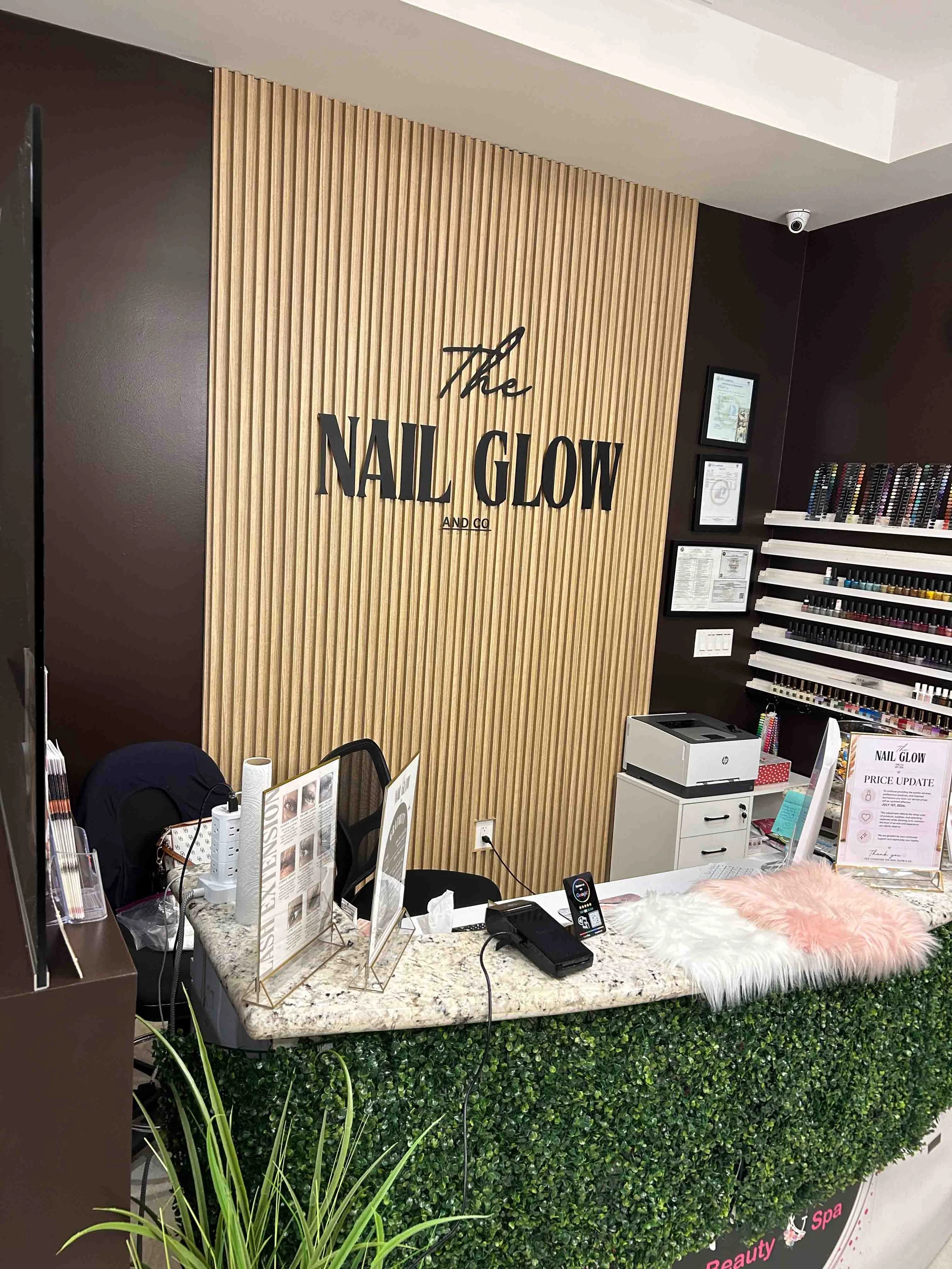 A salon or beauty shop counter with a large green hedge at the front and various items on the marble countertop, including informational signs, a printer, and a makeup brush. There is a sign that reads 'The NAIL GLOW PRICE UPDATE' on the right side of the counter. Behind the counter is a wooden-slatted wall with the words 'The NAIL GLOW AND CO' in black letters, and to the right, a display of nail polish bottles and framed certificates.