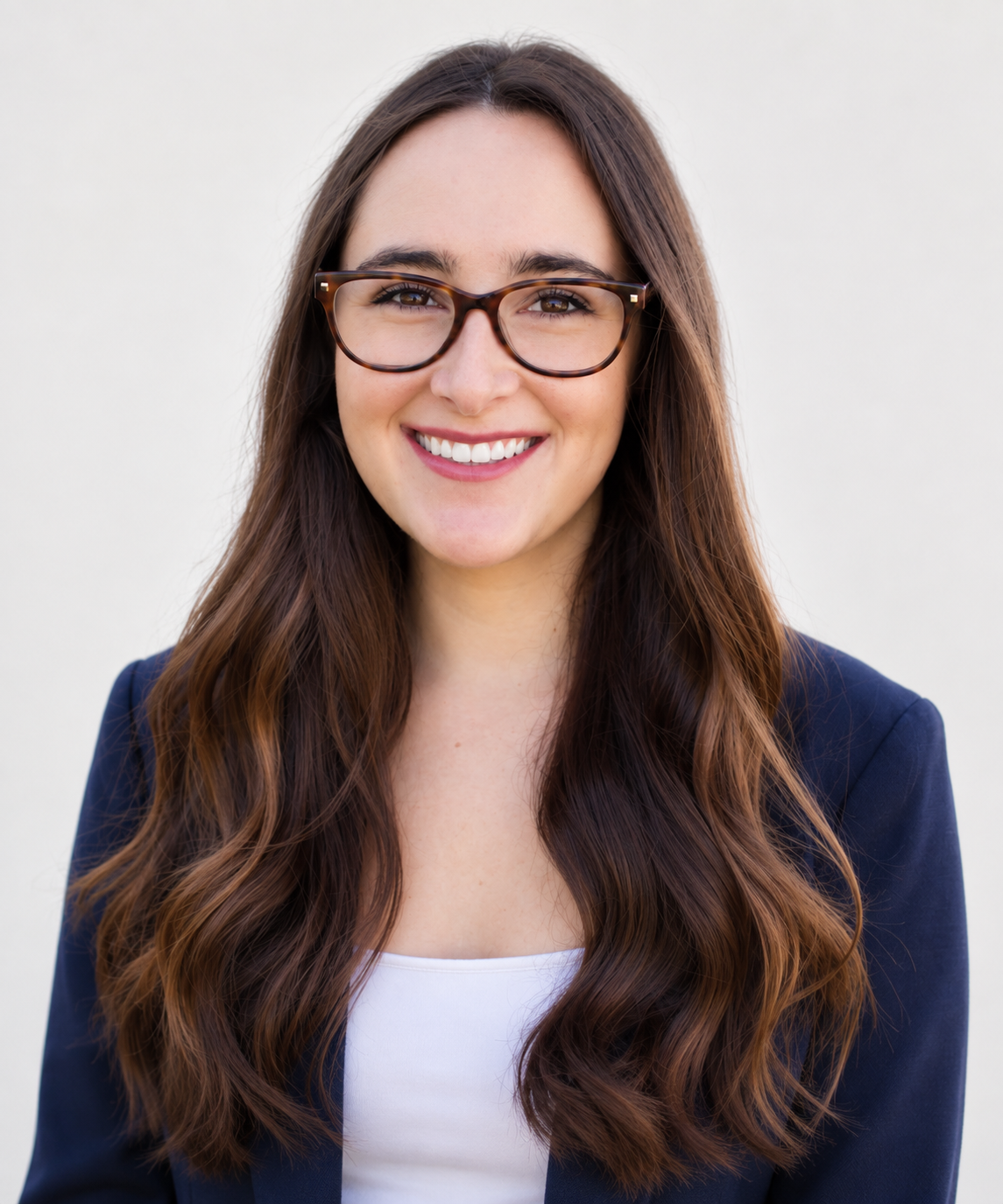 A young woman with long brown hair, wearing glasses and a navy blue blazer, smiling outdoors in a park with green trees and grass in the background.