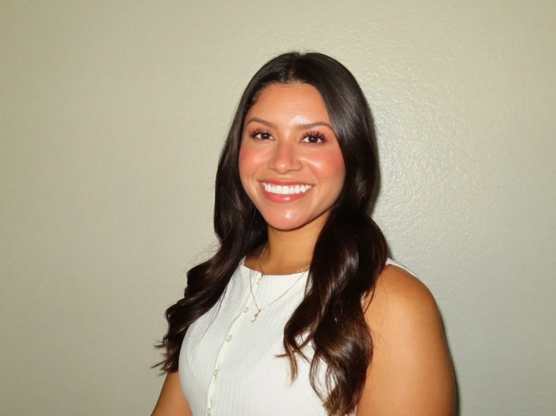 A young woman with long, wavy brown hair, smiling and wearing a white sleeveless top, standing against a plain beige wall.