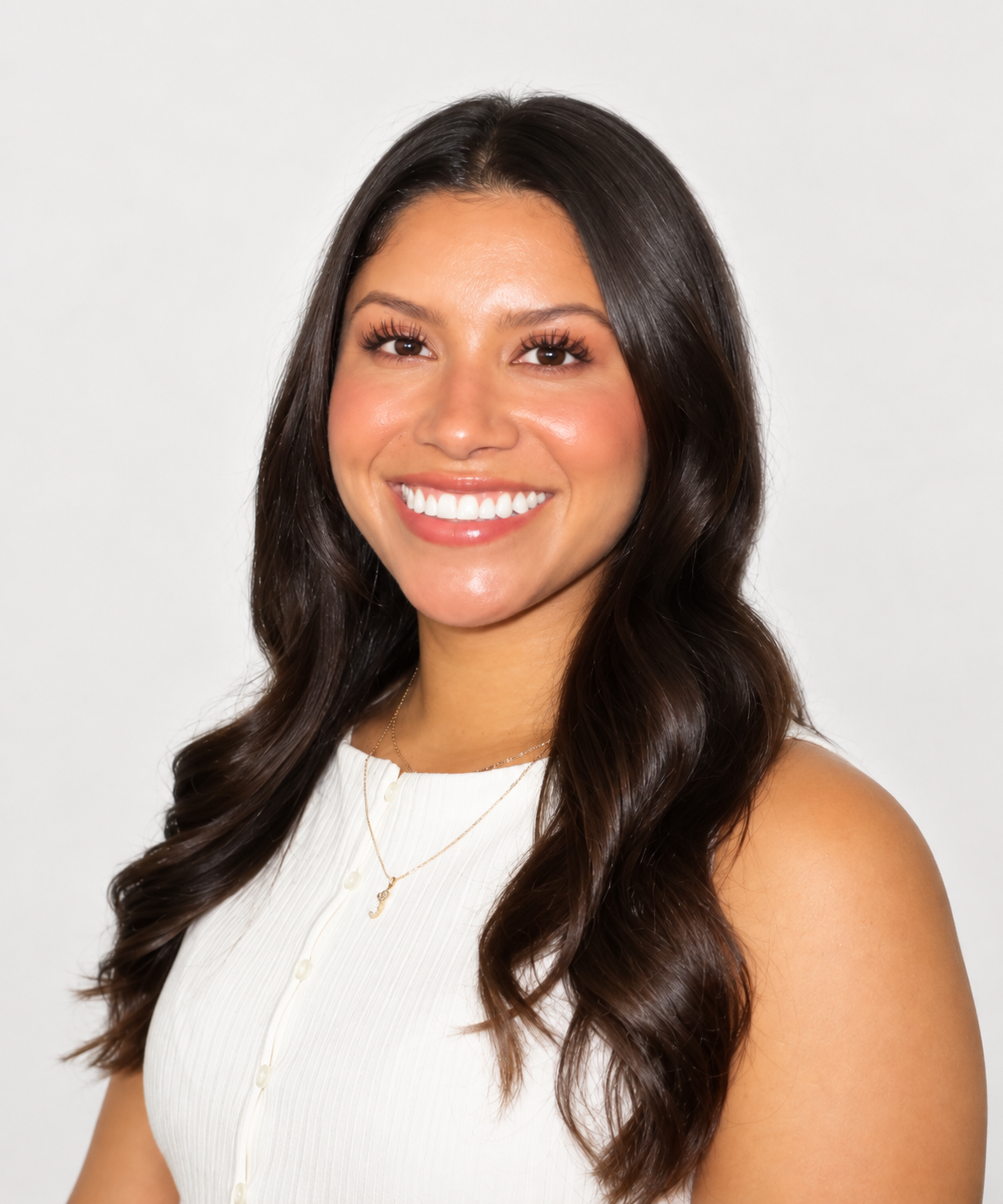 A young woman with long, wavy brown hair, smiling and wearing a white sleeveless top, standing against a plain beige wall.