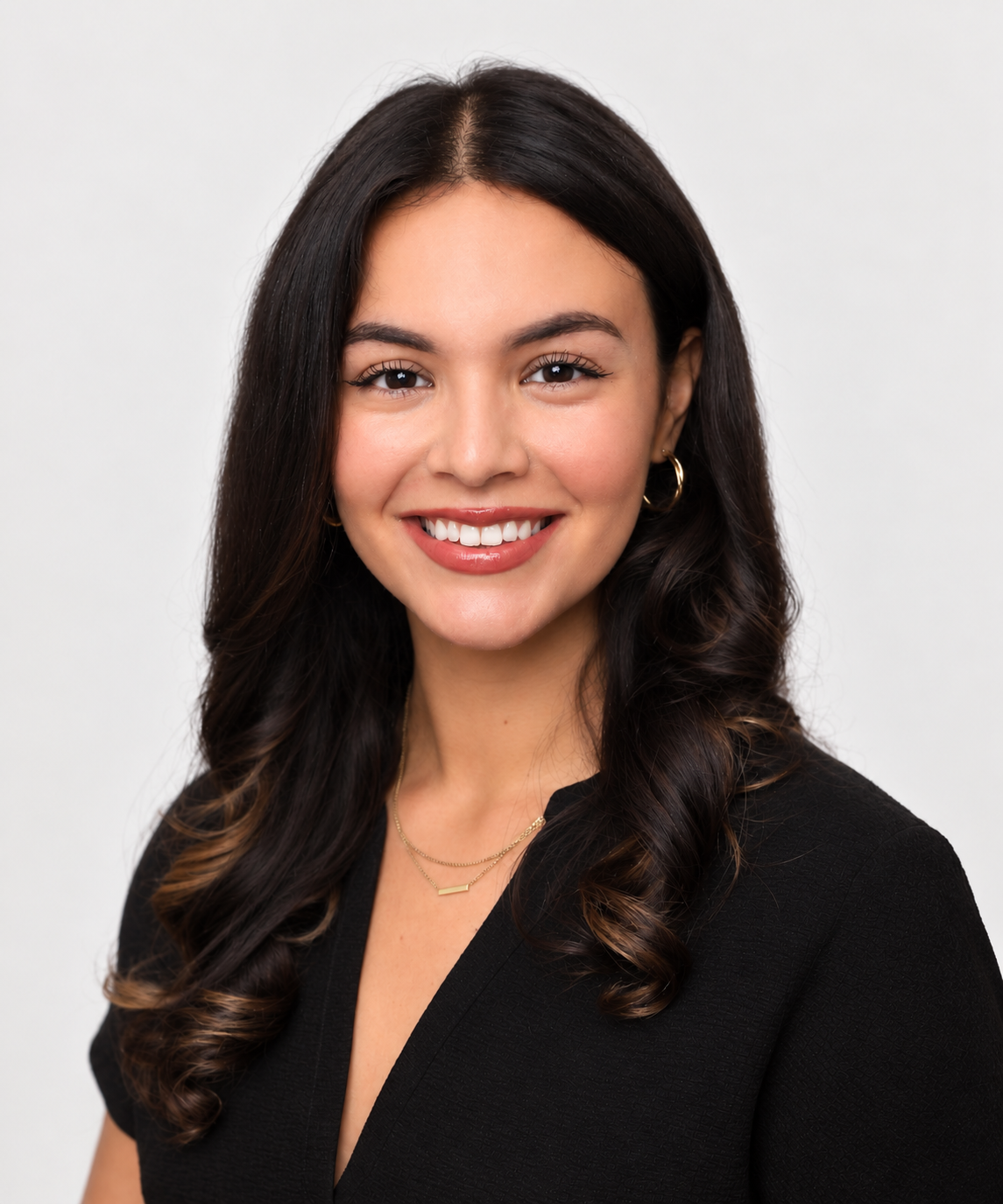 Headshot of a young woman with dark, wavy hair, wearing a black top, gold jewelry, and smiling against a gray background.