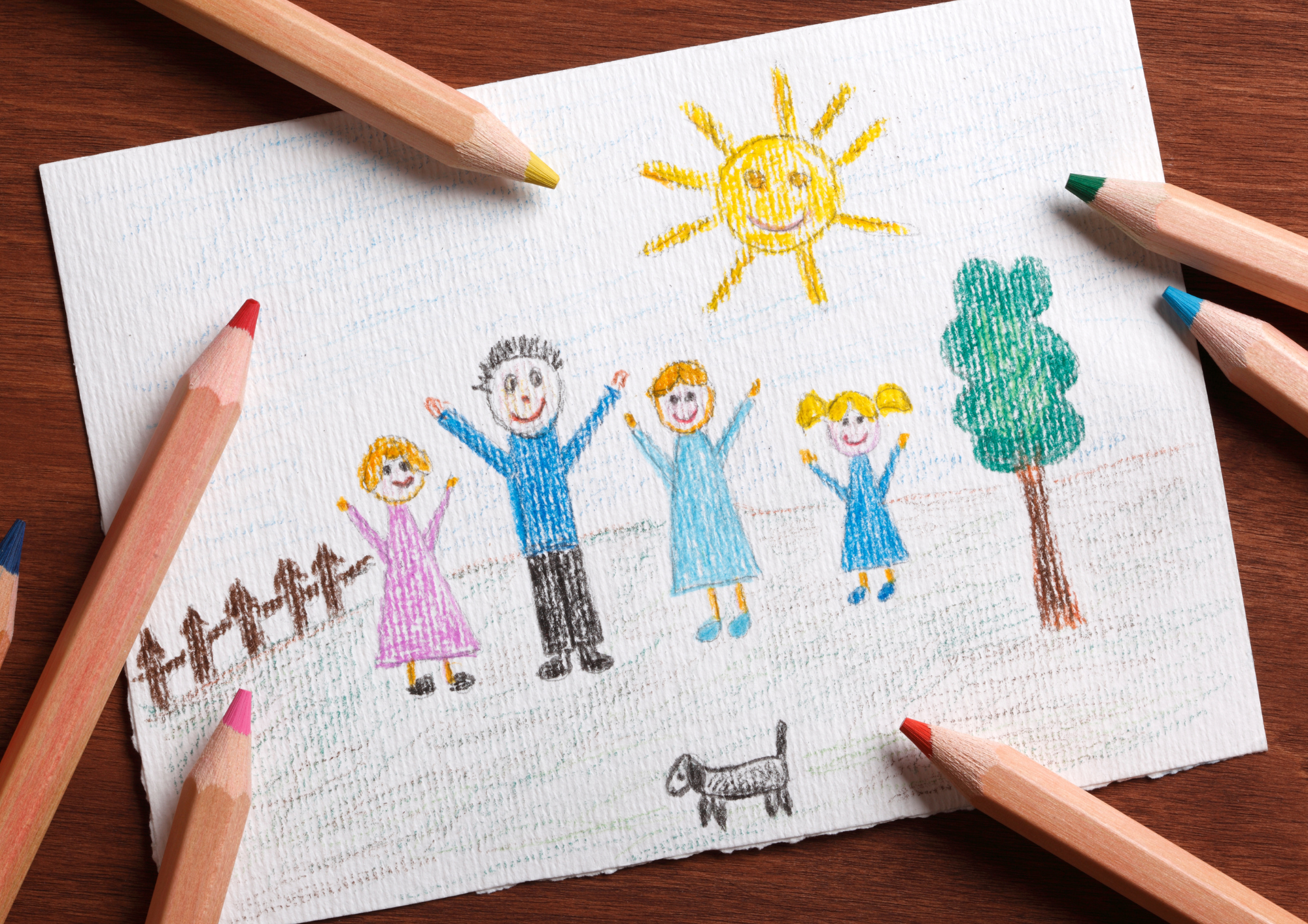 Colorful crayon drawing of a family of four children, a dog, a tree, and a smiling sun in a blue sky on a textured white paper, surrounded by colored pencils.