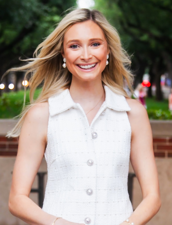 A smiling blonde woman in a white sleeveless dress standing outdoors.