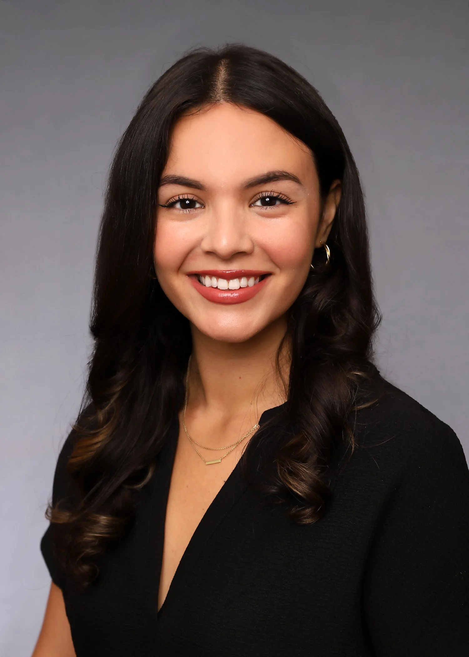 Headshot of a young woman with dark, wavy hair, wearing a black top, gold jewelry, and smiling against a gray background.