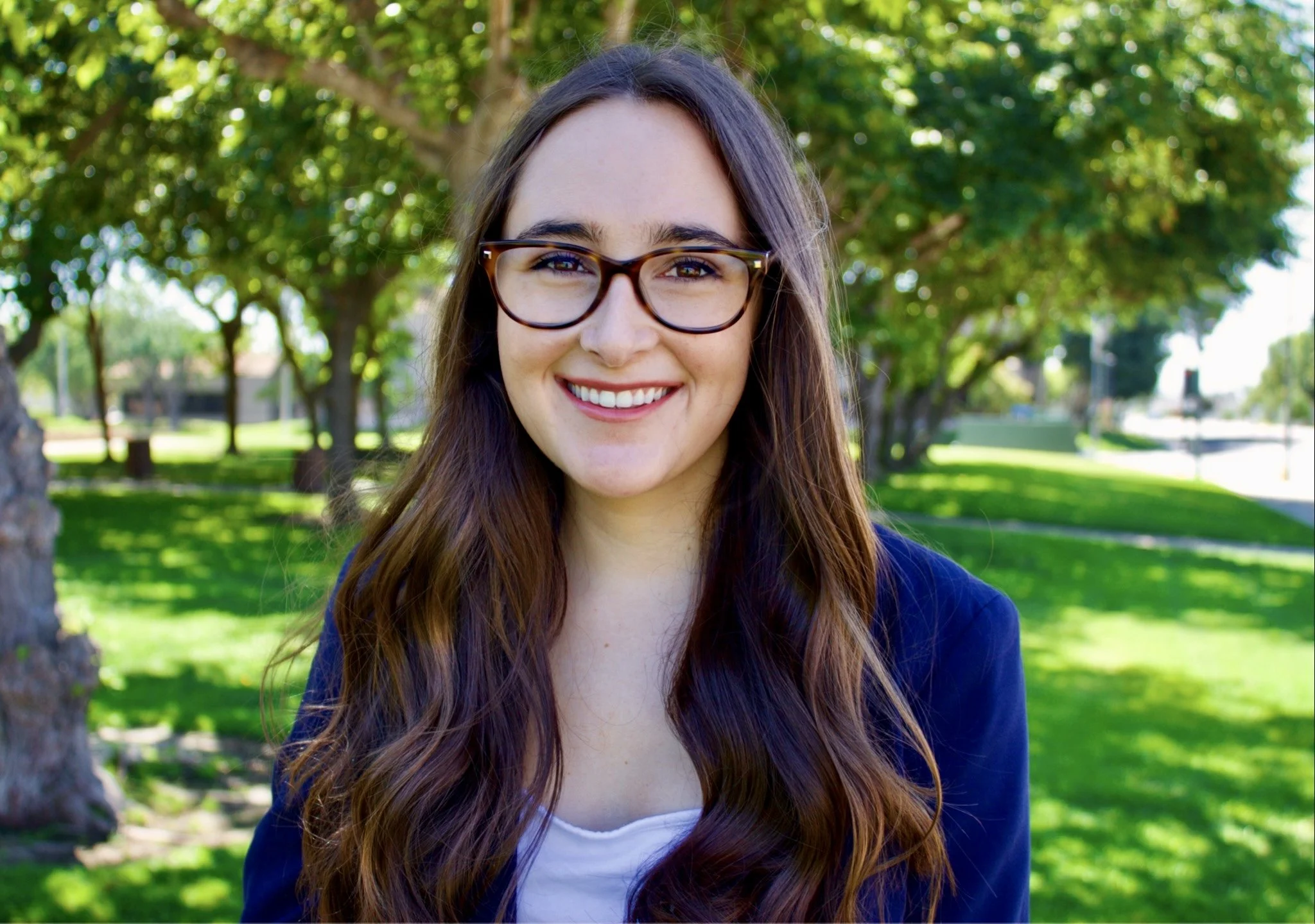 A young woman with long brown hair, wearing glasses and a navy blue blazer, smiling outdoors in a park with green trees and grass in the background.