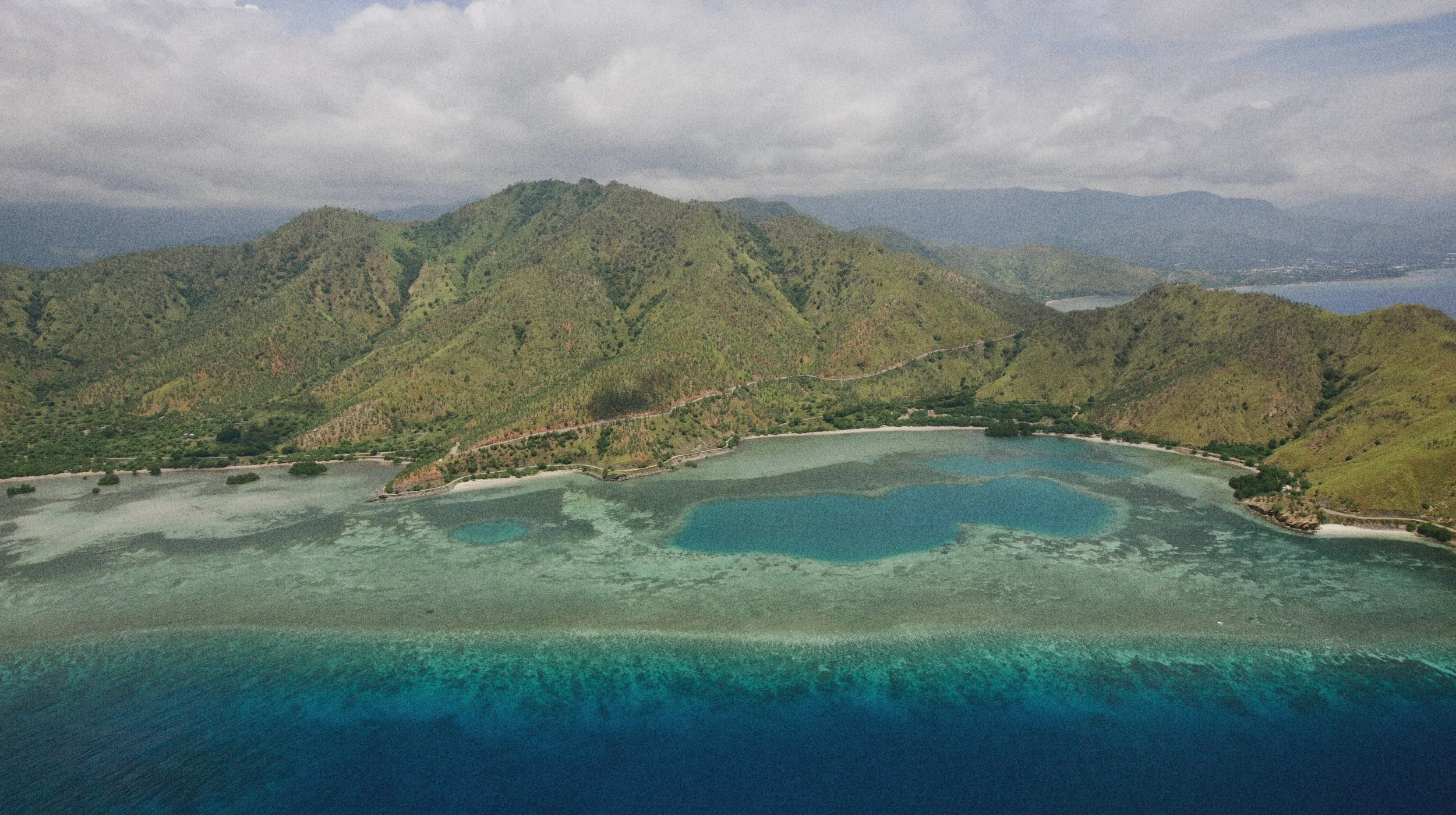 Aerial view of a coastal bay with turquoise water, green hills, and a cloudy sky. Located in Timor-Leste - Premium Indonesian Green Coffee Beans Exporter