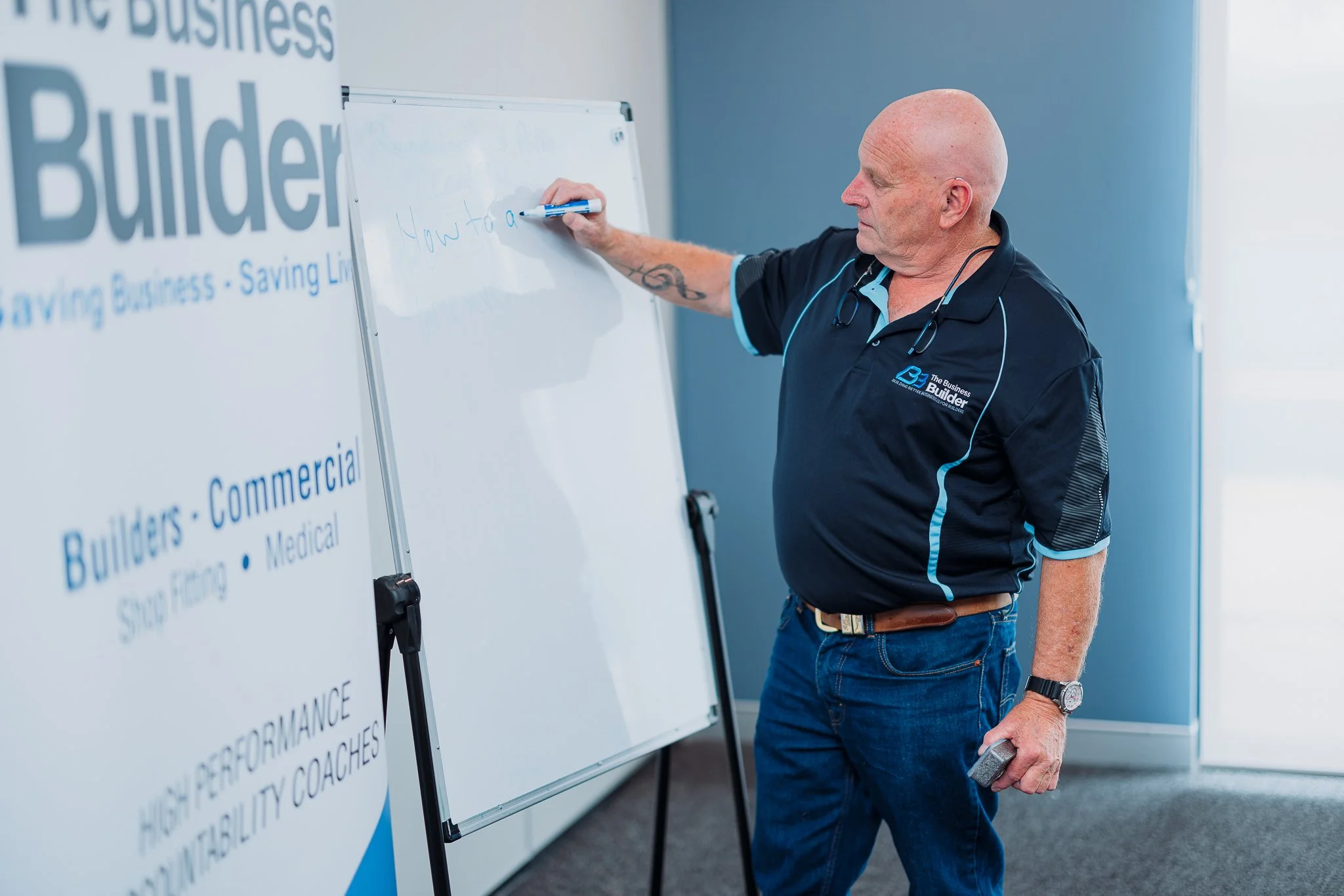 Ian Wholohan, a man writing on a whiteboard during a presentation. The whiteboard has some partially visible text, and there is a poster on the left side with the text 'The Business Builder' and details about business training and coaching.