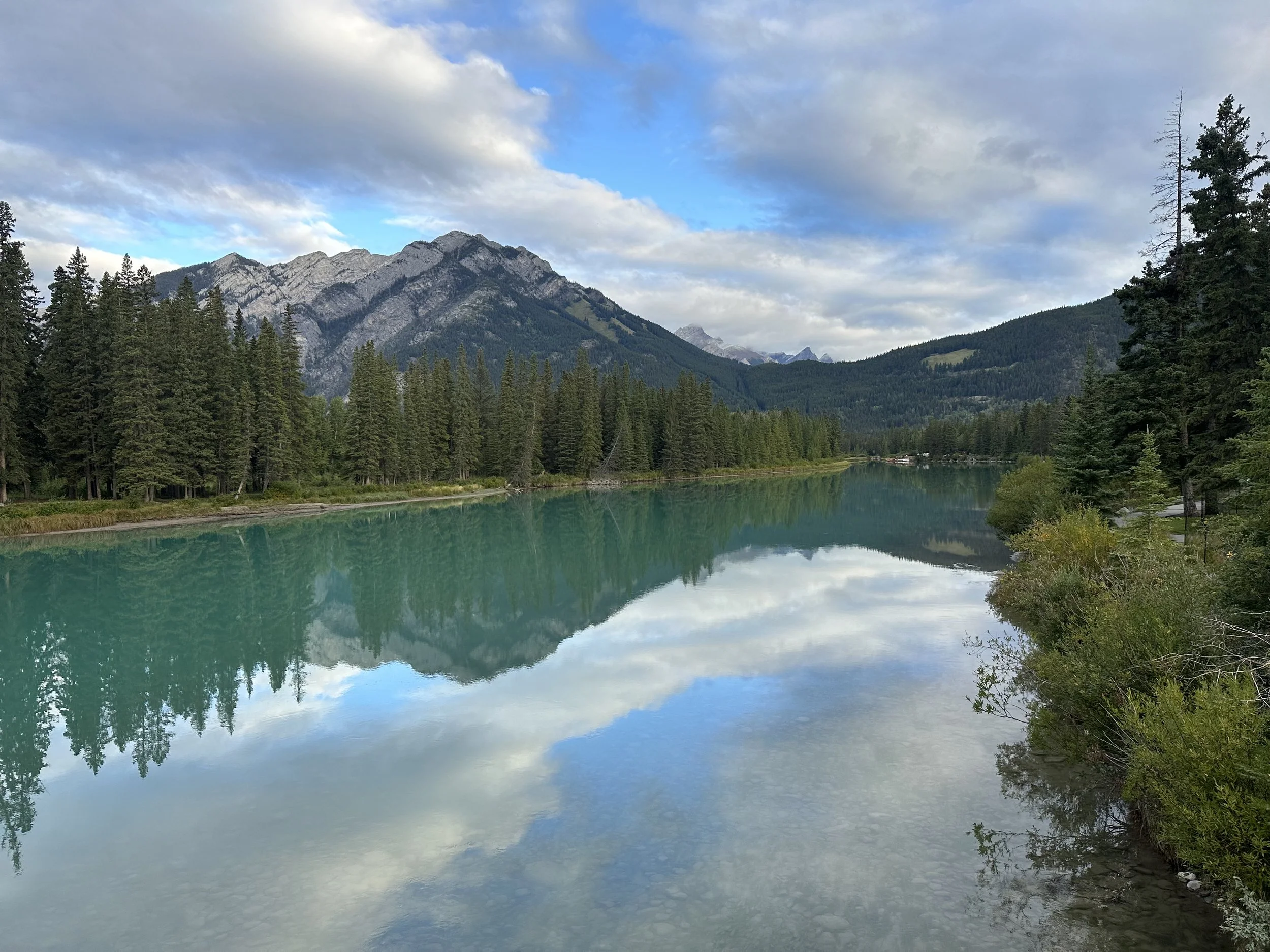 A tranquil mountain lake reflecting the sky in Banff, with pine trees along the shoreline and mountains in the background, the perfect setting for reflection and self-discovery.