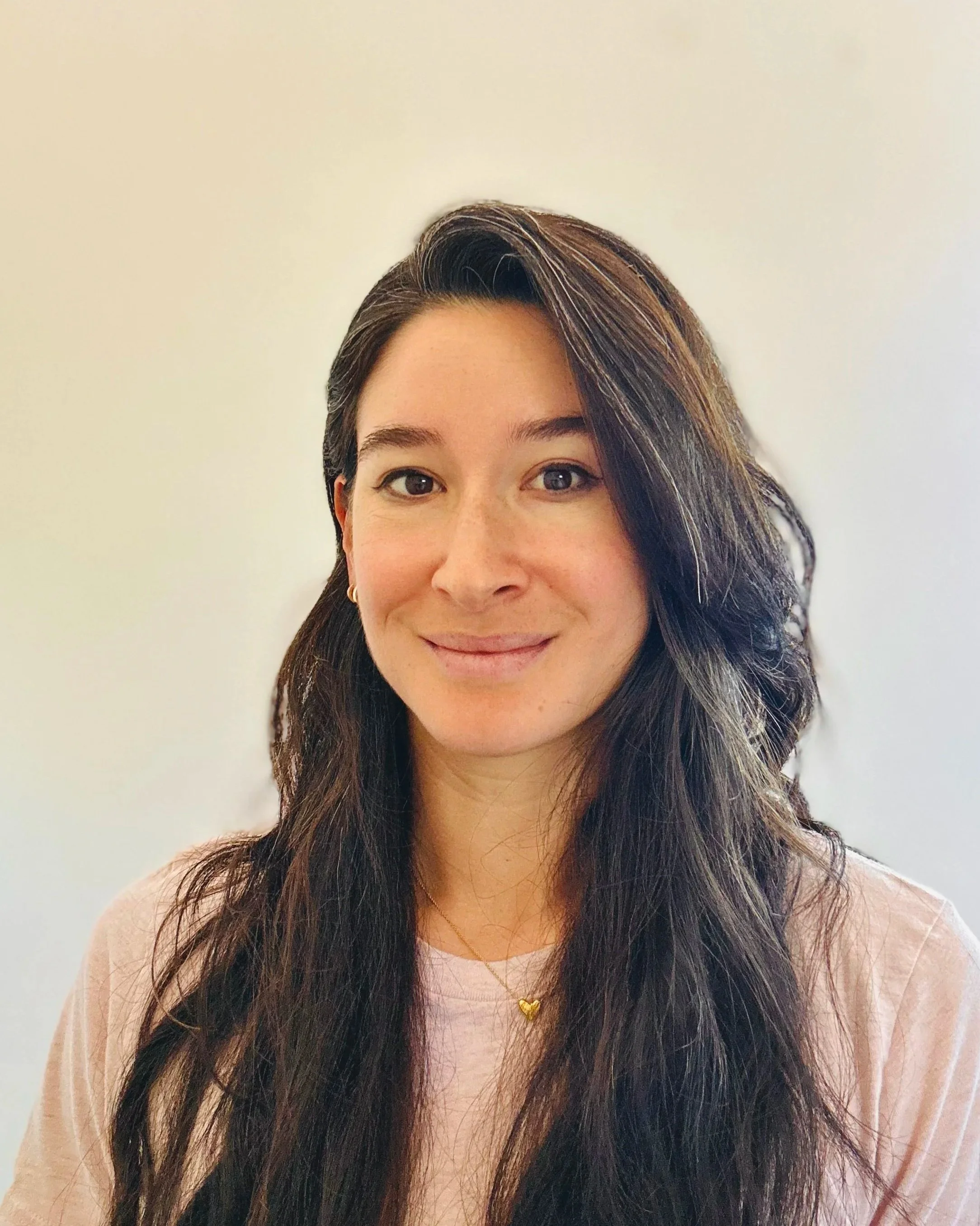 A grounded spiritual woman with friendly and understanding smile, dark brown hair, wearing a light pink shirt and a gold heart necklace, welcoming and inviting deep conversation and self-discovery.
