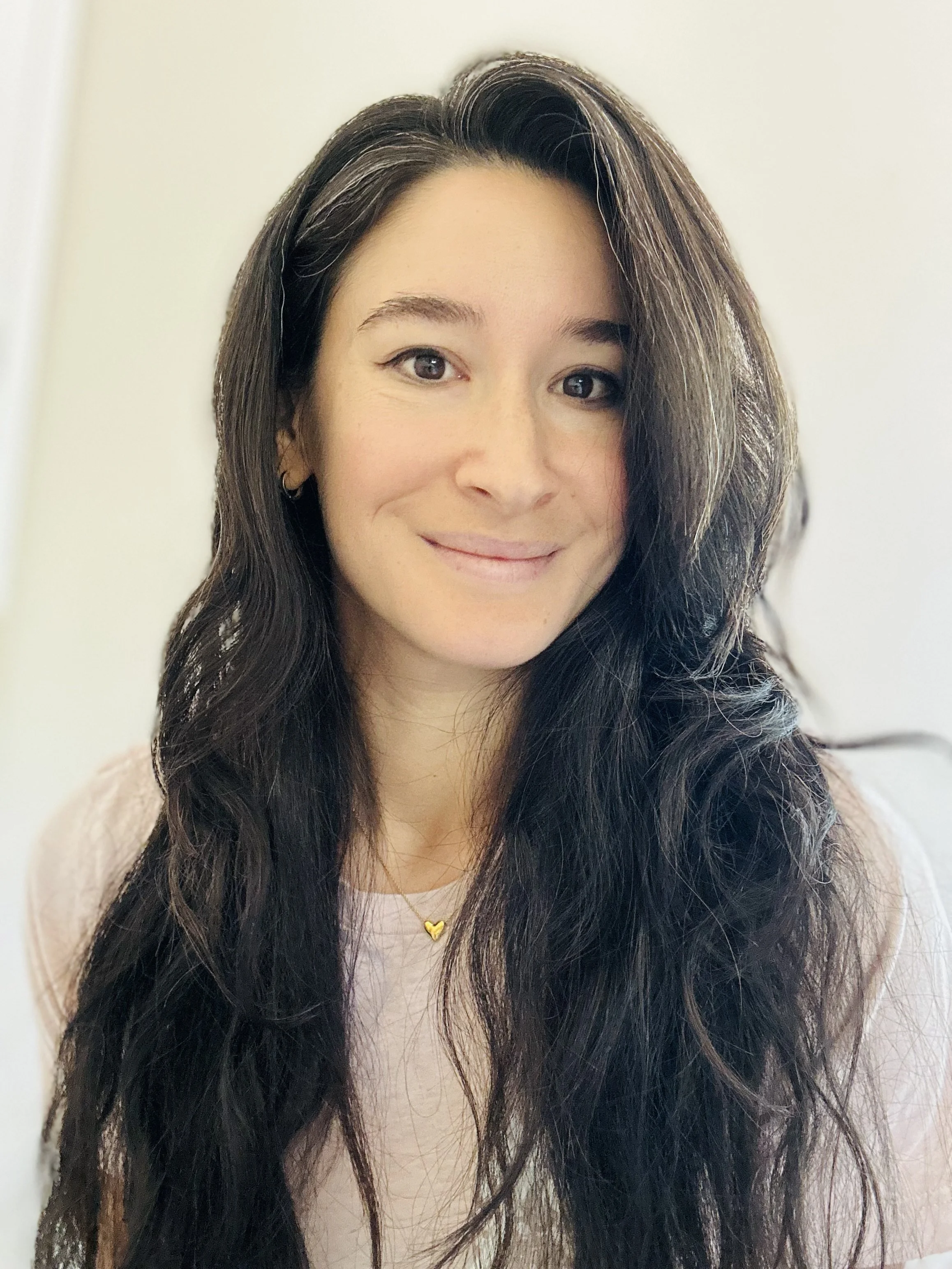 A grounded spiritual woman with friendly and understanding smile, dark brown hair, wearing a light pink shirt and a gold heart necklace, welcoming and inviting deep conversation and self-discovery.