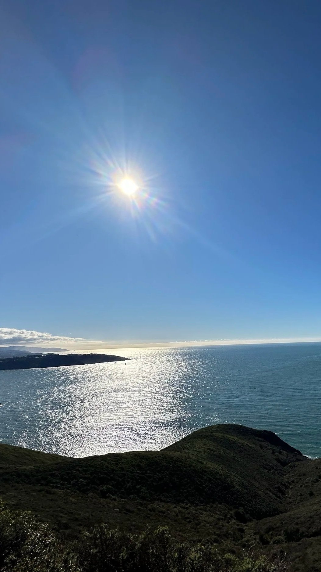 A sunny day over the ocean, with the sun shining brightly in a clear blue sky, reflecting on the water's surface, with green hills in the foreground and distant landforms on the horizon, the perfect setting for reflection and self-discovery.