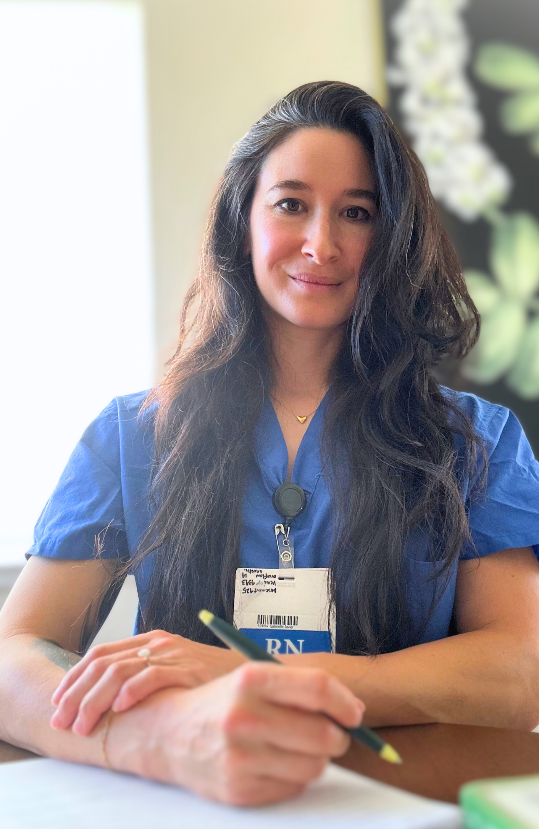 A young, grounded, spiritual woman with friendly and understanding smile, dark brown hair, wearing blue hospital scrubs and a name badge saying "RN," sitting at a table with a pen in her hand, in a welcoming space for doing inner work on self.