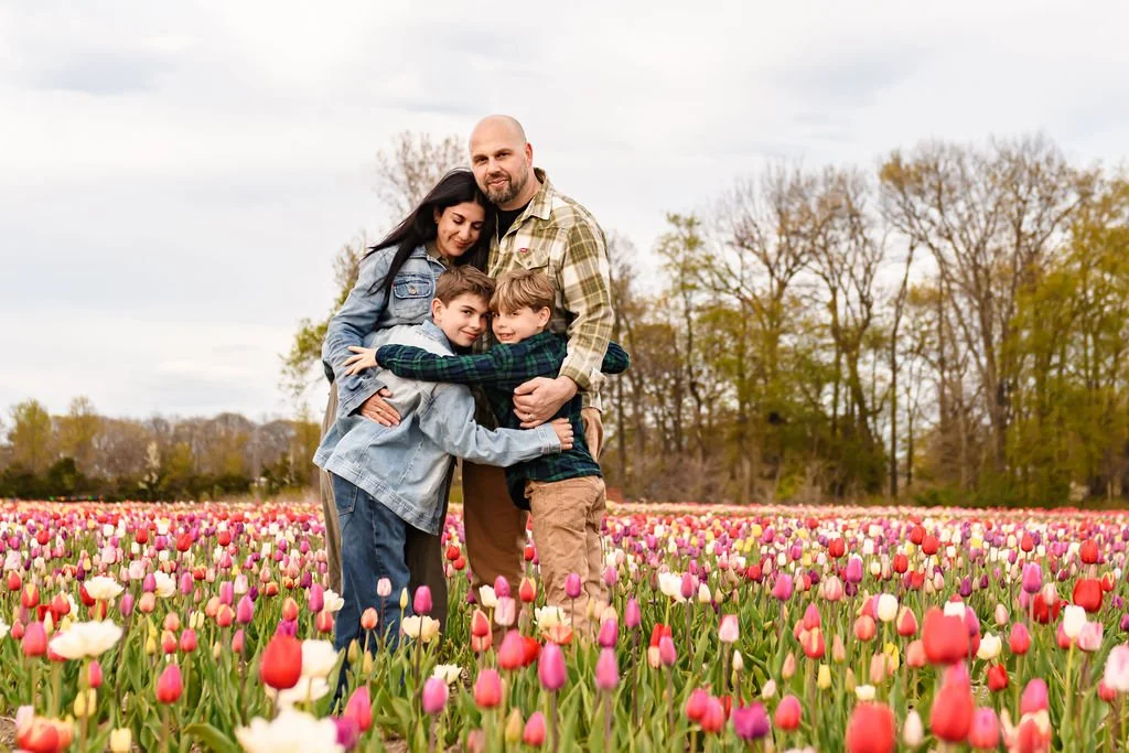 Family of four embracing in a tulip field during daytime, with trees and cloudy sky in the background.