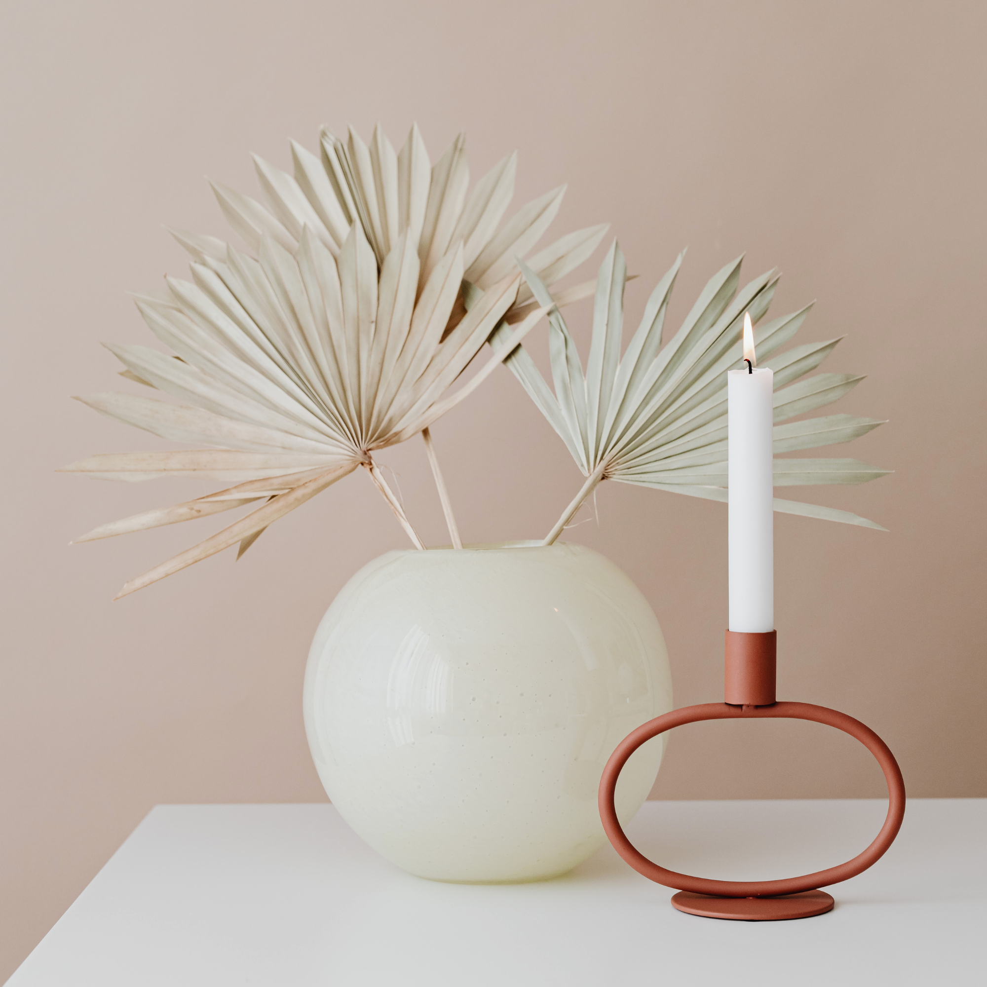 A white vase with dried beige palm leaves and a lit white candle in a terracotta holder on a white surface against a beige background.