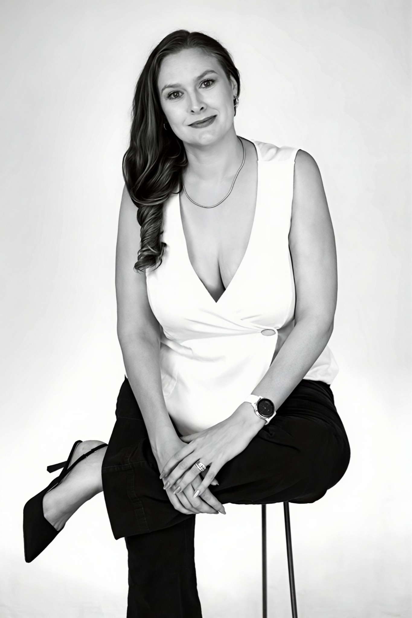Black and whiteigh portrait of a woman sitting on a stool, wearing a sleeveless top, black pants, h heels, and accessories including a watch, rings, earrings, and a necklace, with a slight smile and long wavy hair.