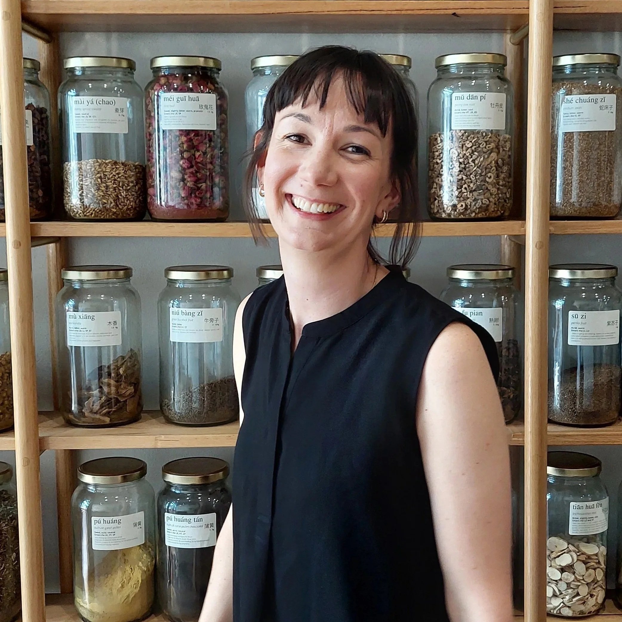 A woman with short dark hair, wearing a black sleeveless top, smiling in front of a wooden shelf filled with glass jars containing various dried herbs and plants.