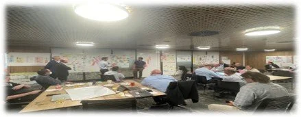 A classroom with students seated at desks and teachers standing at the front, engaging in work.