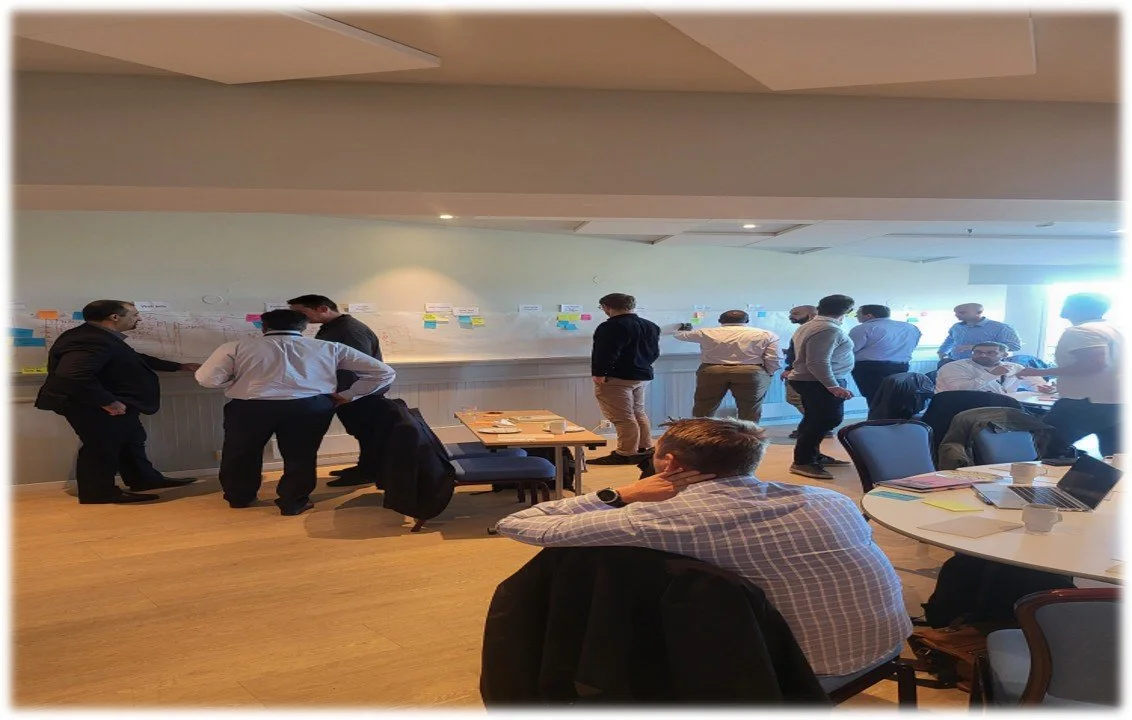 Business professionals in a conference room analyzing a large project timeline on the wall with sticky notes, while some are seated and others stand discussing.