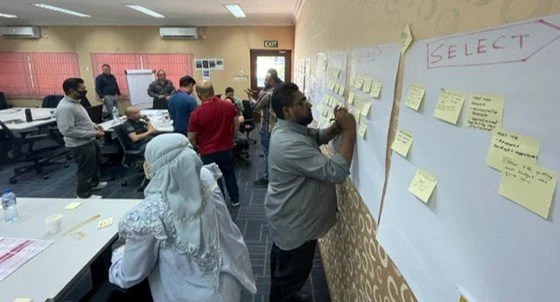 People in a classroom participating in a workshop or training session, with some writing on large paper sheets and sticky notes on the wall, engaging in group activities.