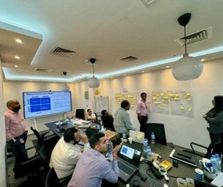 Business meeting in a conference room with people, a large screen, sticky notes on the wall, and laptops on the table.