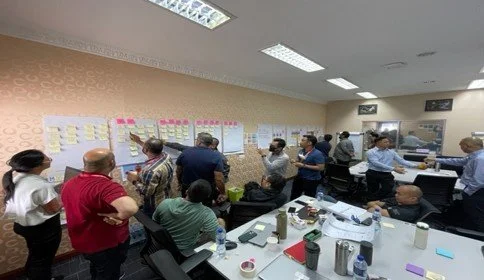 Group of people in a meeting room looking at large papers, posters, or charts on the wall. Some people are standing, some are seated, and they appear to be discussing or analyzing the information displayed.