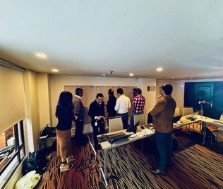 Group of people gathered around a table with laptops in a conference room with carpeted floors and a large flat-screen TV.