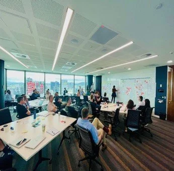People attending a meeting or presentation in a modern conference room with large windows and a digital display screen.