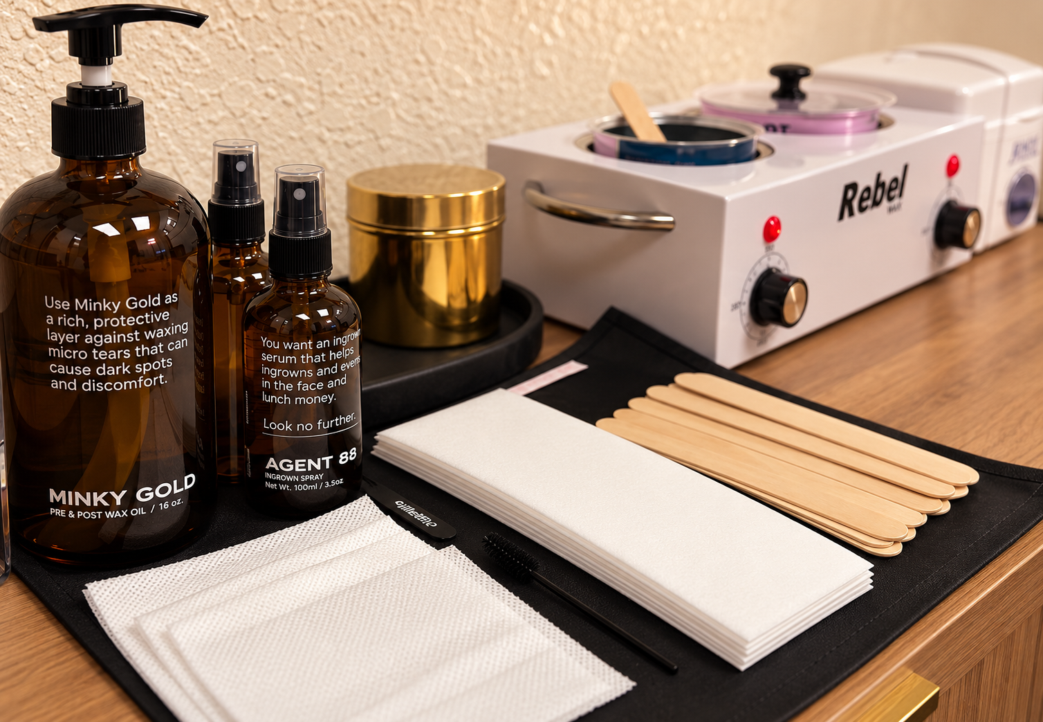 Setup of waxing supplies including bottles labeled for waxing products, wooden spatulas, strips, a black tray, and a wax warmer on a wooden table.