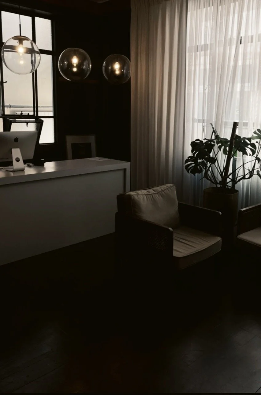 Dimly lit office or study with a modern desk, computer, beige armchair, large potted plant, window with sheer curtains, and hanging glass globe lights.