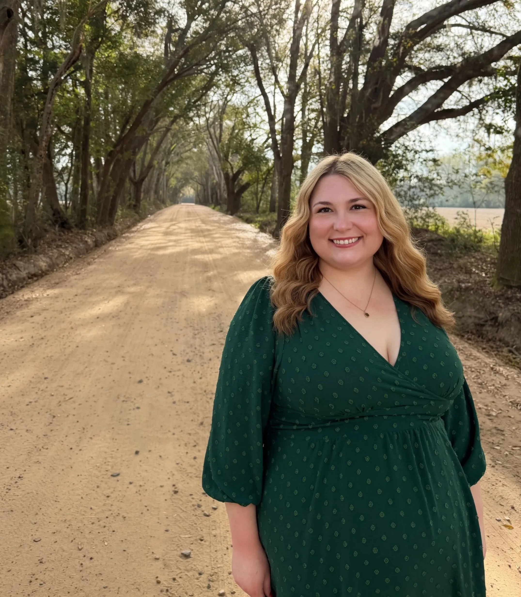 A woman with blonde hair in a green dress standing on a dirt road lined with trees.